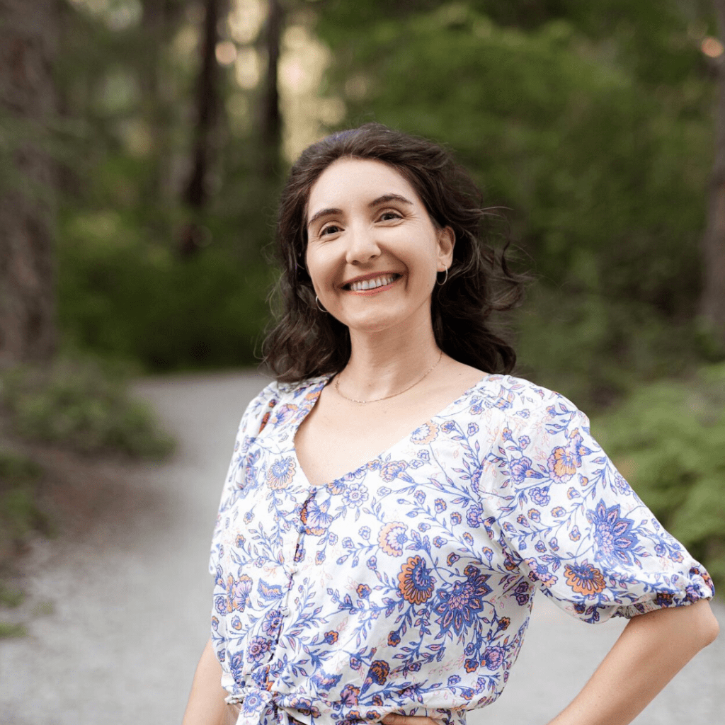 Kimberly Absher outside in the woods in a shirt with flowers on it, brown hair, and one hand on her hip
